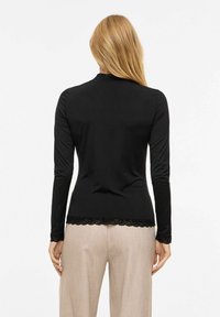 Svart langermet topp med blondekant, med tettsittende design og glatt stoff. Kombinert med lyse beige bukser.