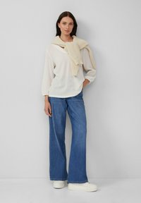 Vrouw met donker haar, gekleed in een witte blouse, een crèmekleurige trui over haar schouders gedrapeerd, blauwe wijde jeans en witte platform sneakers.
