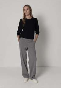 Czarna koszula z długim rękawem z białą lamówką, szare szerokie spodnie w paski oraz białe trampki. Model stoi pewnie, mając ręce w kieszeniach.