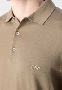 Polo en maille beige avec col, deux boutons et "BOSS" brodé sur le côté gauche de la poitrine, porté par une personne.