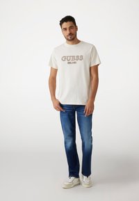 Bijela pamučna majica s natpisom "GUESS EST. 1981" u smeđim slovima, kombinirana s plavim trapericama i bijelim tenisicama. Jednostavan, ležeran dizajn.