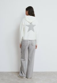 Sudadera con capucha blanca recortada con patrón de estrellas grises en la parte trasera, combinada con vaqueros de pierna ancha gris claro con detalles de estrellas. Tela suave, estilo casual.