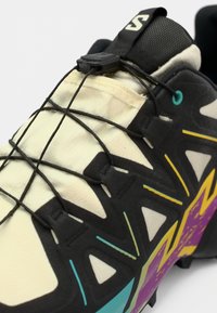 Chaussure de sport noire et crème avec une tige texturée, un système de laçage en cordon élastique et des motifs géométriques colorés en turquoise, violet et jaune.