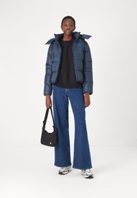 Marine pufferjack met een ritssluiting en afneembare capuchon, gecombineerd met een zwarte sweatshirt, brede denim jeans en een zwarte handtas. Zilveren sneakers.