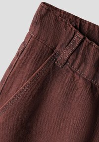 Primo piano di un tessuto denim marrone che mostra la vita, il passante per la cintura, la cucitura e i dettagli delle cuciture di un indumento.
