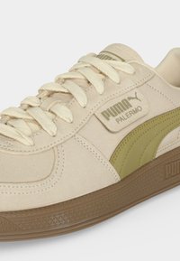 Μπέζ αθλητικό παπούτσι Puma Palermo με κρεμ κορδόνια, ελιά πράσινη πλευρική ραφή και καφέ υφασμένη σόλα σε λευκό φόντο.