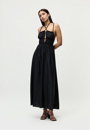 Kobieta w długiej czarnej sukni na szyję z wycięciami i sandałach na obcasie, stojąca na tle jasnego, jednolitego tła.