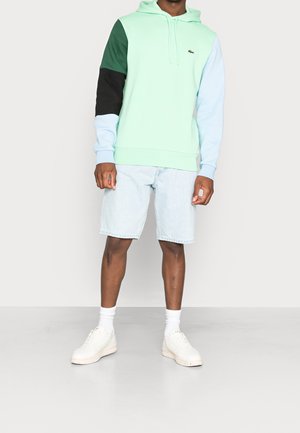 Homme portant un sweat à capuche bicolore avec des manches vertes, noires et bleu clair, un short en denim clair, des chaussettes blanches et des baskets blanches.
