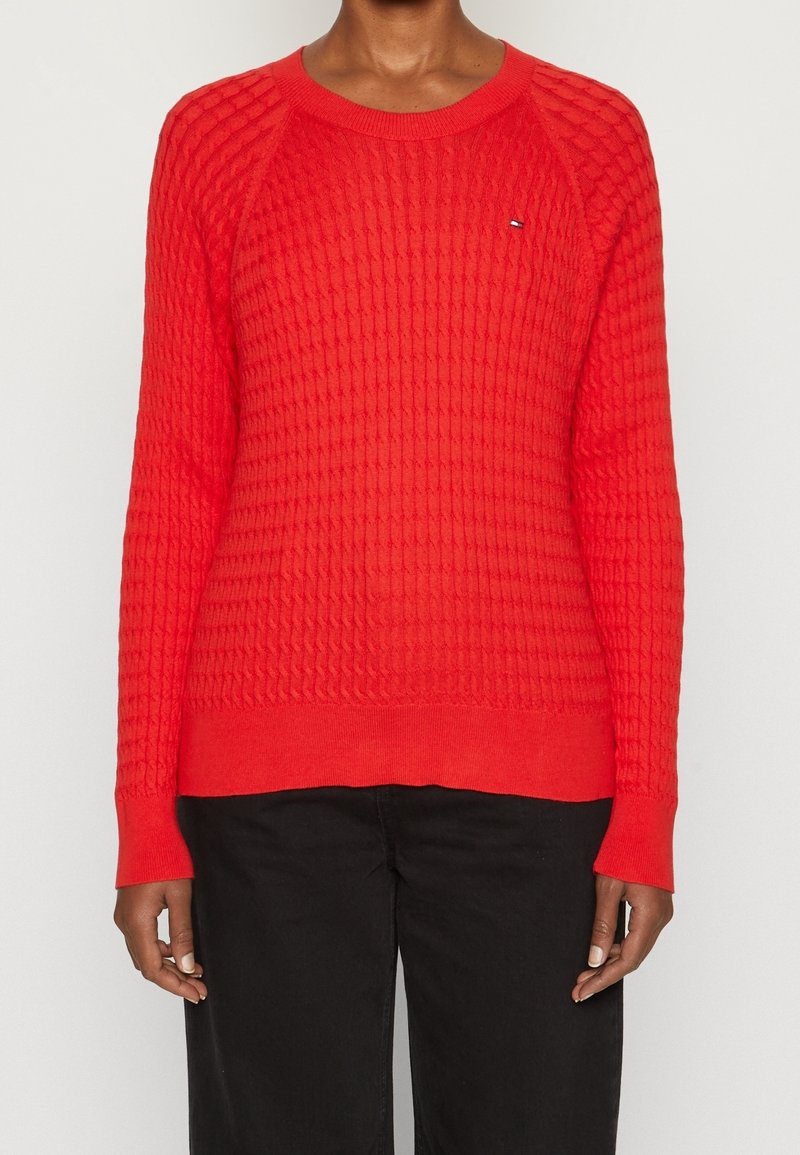 Personne portant un pull tricoté rouge vif à texture avec des manches longues et un pantalon noir, debout devant un fond clair uni.