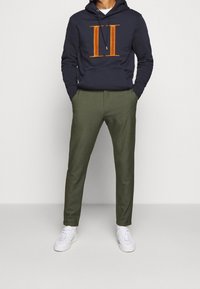 Marinblå hoodie med orange sammet "II"-logotyp, kombinerad med olivgröna skräddarsydda byxor och vita sneakers, har en avslappnad passform och justerbar huva.