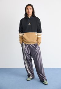 Črna in rjava kapuca s tremi belimi črtami na rokavih, v kombinaciji z sijočimi sivimi trenirkami, ki imajo bele stranske črte in Adidasov logotip.