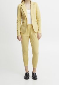 Blazer jaune en mélange de coton avec deux poches avant, associé à un pantalon de jogging à cordon assorti. Débardeur blanc en dessous, chaussures noires.