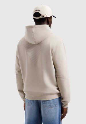Mann trägt beige Hoodie mit strukturiertem dreieckigem Muster auf dem Rücken und hellblauen Jeans, von hinten zu sehen, trägt zudem eine cremefarbene Baseballkappe.