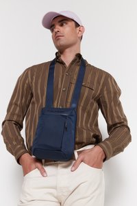 Borsa a tracolla navy con tessuto testurizzato, tracolla regolabile e branding discreto. Indossata sopra una camicia a righe marroni e pantaloni color crema.