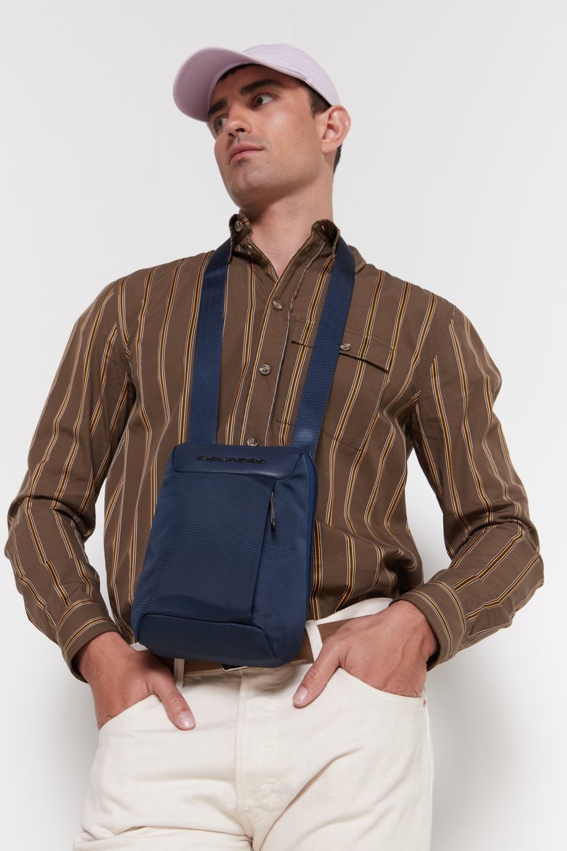 Borsa a tracolla navy con tessuto testurizzato, tracolla regolabile e branding discreto. Indossata sopra una camicia a righe marroni e pantaloni color crema.