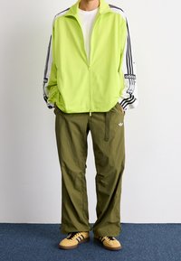 Lime green zip-up jacket with white and black sleeves, paired with loose olive green pants and yellow shoes with black stripes.