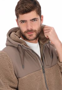 Sudadera con capucha marrón con cremallera, textura suave de forro polar, parte superior lisa en contraste, cordones ajustables y corte holgado.