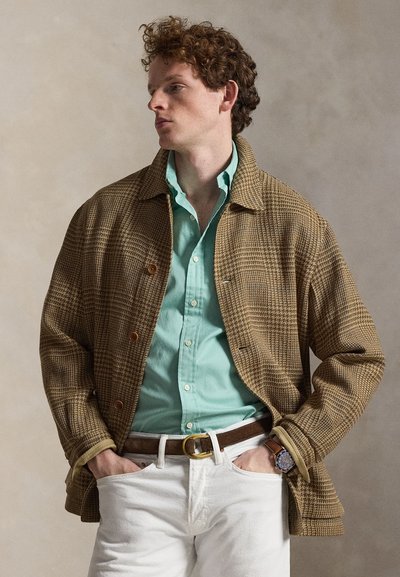 Άνδρας με σγουρά μαλλιά που φοράει καφέ καρό σακάκι, ανοιχτό μπλε πουκάμισο με κουμπιά, λευκό παντελόνι, καφέ ζώνη και ρολόι, με τα χέρια στις τσέπες.