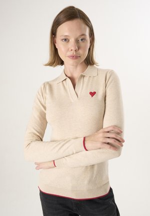 Donna con capelli castano chiaro che indossa un maglione beige a manica lunga con toppa a forma di cuore rossa e rifiniture rosse, in piedi con le braccia incrociate davanti a uno sfondo semplice.