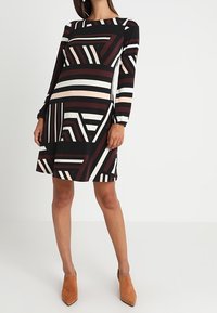 Long-sleeve dress in black with geometric stripes in beige, white, and burgundy. Available with a round neckline and slightly flared bottom.