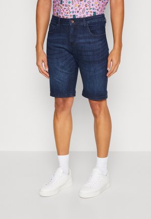 Persoon draagt donkerblauwe denim shorts, witte sokken, witte sneakers en een roze shirt met kleurrijke kleine prints, staand tegen een effen achtergrond.
