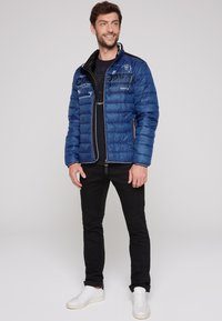 Giacca a vento blue con texture a quadri, dettagli neri e zip. Indossata sopra una t-shirt nera con grafiche, abbinata a pantaloni neri e sneaker bianche.