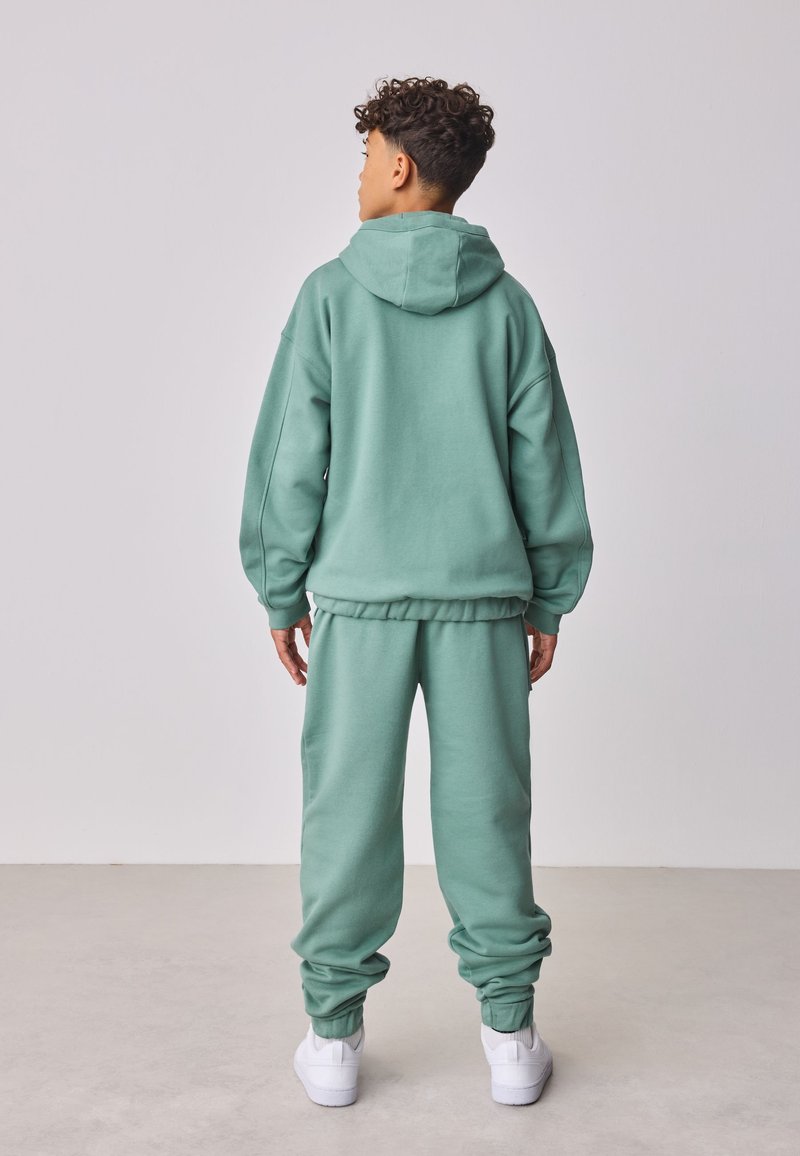 Junge mit lockigem Haar steht mit dem Rücken zur Kamera, trägt einen grünen Hoodie, passende Jogginghose und weiße Sneakers auf einfarbigem Hintergrund.