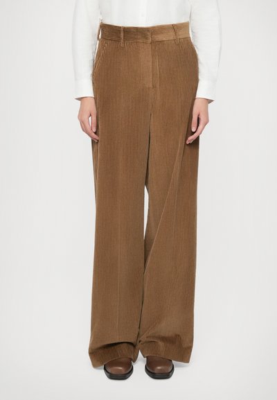 Pantaloni corduroy larghi marroni indossati con una camicia bianca a maniche lunghe e scarpe marroni su uno sfondo semplice.