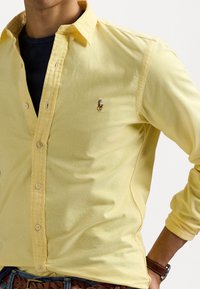 Żółta koszula zapinana na guziki z teksturowanej tkaniny, z małym wyszywanym logo i klasycznym kołnierzem. Noszona na granatowej koszulce.