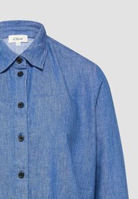 Blaues Denimhemd mit schwarzen Knöpfen und klassischem Kragen, teilweise dargestellt vor einem schlichten hellgrauen Hintergrund.