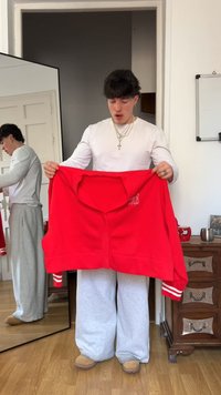 Sweatshirt vermelho cortado com ajuste oversized, punhos canelados e abertura frontal. Modelo usando calças de moletom cinzas e pantufas beges. Espelho ao fundo.