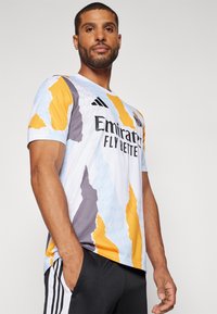 Maglia sportiva bianca con motivi astratti blu, arancioni e grigi. Presenta il testo nero "Emirates Fly Better" e il logo Adidas. Maniche corte.