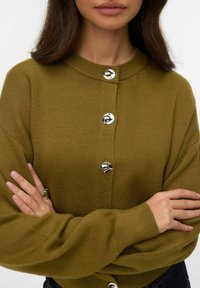 Oliwkowo zielony sweter na drutach z zaokrąglonym dekoltem, pięcioma błyszczącymi srebrnymi guzikami oraz długimi, luźnymi rękawami. Gładka faktura.