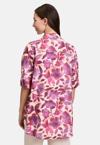 Kwiatowa bluzka z kremowym tłem i dużymi fioletowymi oraz różowymi wzorami kwiatowymi, z krótkimi rękawami i luźnym krojem. Przedstawiony widok z tyłu.