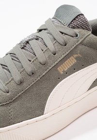 Sneaker in suede grigio con una striscia laterale bianca, lacci testurizzati e logo Puma dorato sul lato. Materiale morbido e design slanciato.