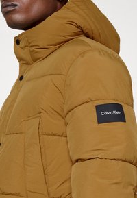 Bruine gewatteerde jas met een capuchon, grote frontzak en zwart Calvin Klein-logo op de mouw. Textuurstof met zichtbare stiksels.