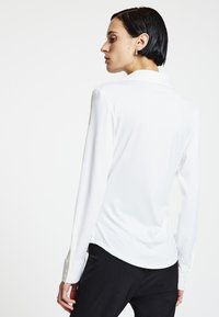 Weiße Bluse mit langen Ärmeln, glatter, glänzender Textur, taillierter Schnitt und klassischem Kragen. Mit geknöpften Manschetten und Seitenschlitzen.