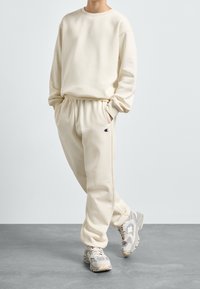 Krémová mikina a odpovídající tepláky vyrobené z měkké látky. Mikina má kulatý výstřih; tepláky mají elastický pas a logo.