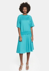 Turkusowa sukienka z miękkiego materiału o luźnym kroju. Posiada krótkie rękawy, talię ściągniętą w pasie oraz rozkloszowaną spódnicę. Srebrne sandały dopełniają całość.