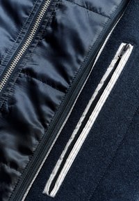 Tejido azul marino con textura acolchada, que presenta una cremallera de metal plateado y un bolsillo frontal con cremallera blanca en contraste.