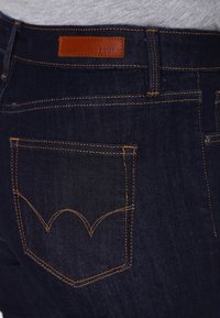 Mörkblå denimjeans med kontrasterande orange sömmar, läderlabel på midjan och bakfickan prydd med en distinkt bågdesign.