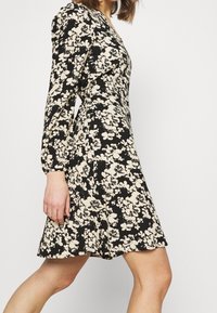 Svart klänning med ett blommönster i krämfärg. Långa puffärmar och en figurnära bodice som leder till en utsvängd kjol. Slät tyg med subtil textur.