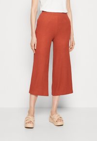 Femme portant un pantalon large court de couleur rouille et des sandales à plateforme beige, debout devant un fond blanc.