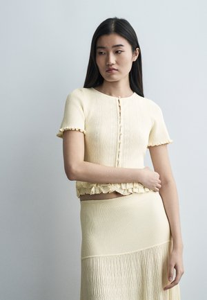 Jeune femme aux longs cheveux bruns portant un haut boutonné à manches courtes jaune pâle et une jupe côtelée assortie, sur un fond uni.