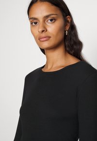 Must värviline, soonikkoes pikkade varrukatega topp, millel on ümmargune kaelus. Kude näeb välja pehme ja pisut tekstureeritud, rõhutades vormi jälgivat siluetti.