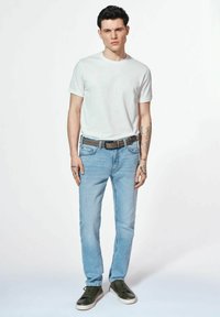 Jasnoniebieskie dżinsy, biała koszulka z okrągłym dekoltem, brązowy pasek i zielone sneakersy. Tatuaże widoczne na ramionach. Stoi na jednolitym tle.