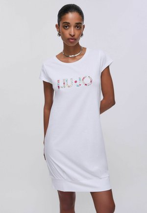 Donna che indossa un abito bianco a maniche corte con logo colorato "LIU•JO", collana d'argento e orecchini a cerchio in argento, in piedi davanti a uno sfondo semplice.
