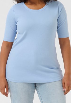 Kvinde iført en lyseblå ribbet t-shirt med korte ærmer og lyseblå jeans, stående med armene afslappet ned langs siderne.
