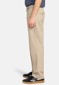 Chinos beige à coupe droite, en tissu de coton. Caractéristiques : poches latérales et une poche arrière avec un bouton, associés à des baskets noires.