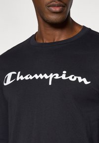 Maglietta nera in cotone con un grande logo "Champion" bianco sul davanti, con scollo rotondo e maniche corte.